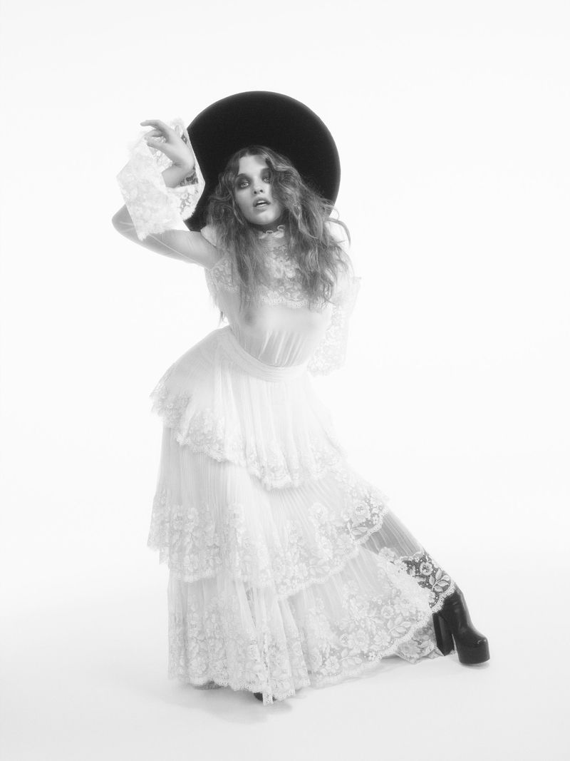 Black and white portrait of a woman in a dress and large hat posing with raised arms in a studio setting with bright, even lighting.
