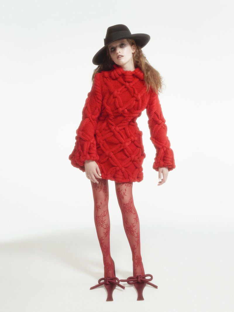 Fashionable young adult woman in a red knit dress and red tights poses in a studio wearing a wide-brimmed black hat and red bow heels.