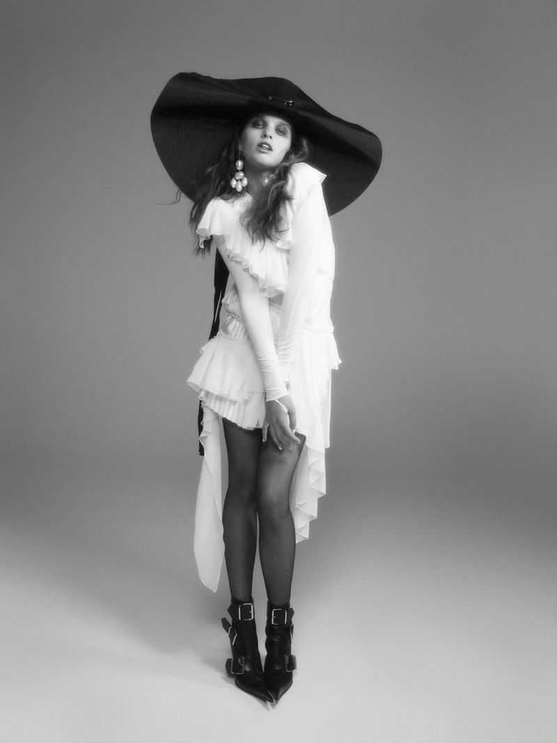 Ever Anderson wearing a large black hat and ruffled white dress against a seamless grey backdrop