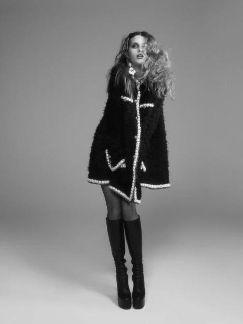 Full-length shot of a fashion model with wavy hair in a black coat and black platform boots, set against a neutral gray background.