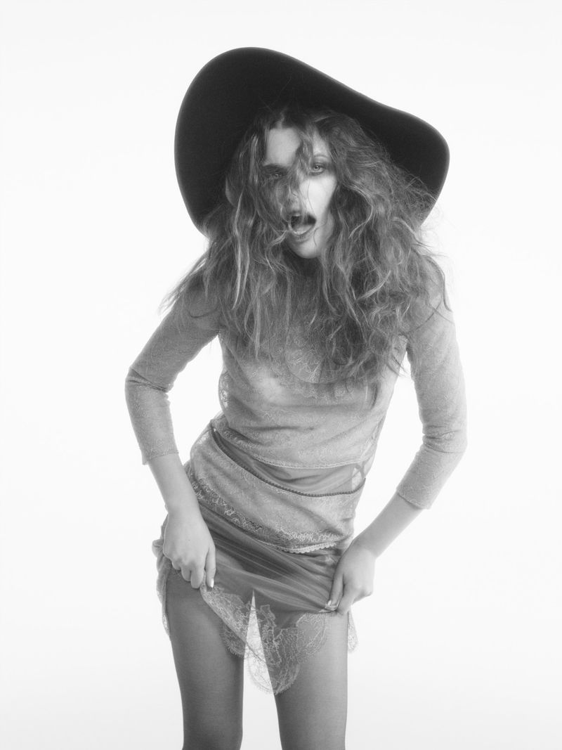Monochrome portrait of a model in a dark hat and semi-sheer dress, dynamically posed against a stark white backdrop.