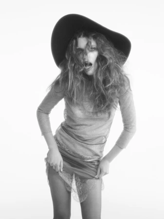 Monochrome portrait of a model in a dark hat and semi-sheer dress, dynamically posed against a stark white backdrop.