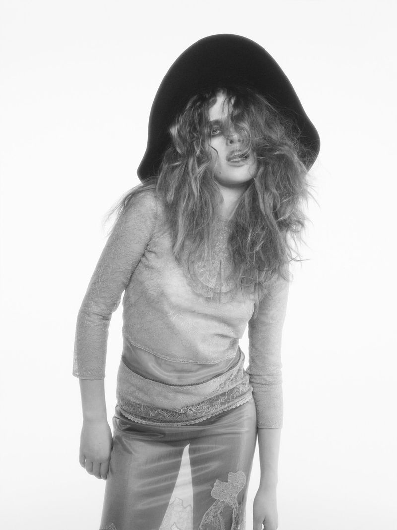Black and white portrait of a woman with long wavy hair wearing a large hat, a long-sleeved top, and a skirt against a plain white background.