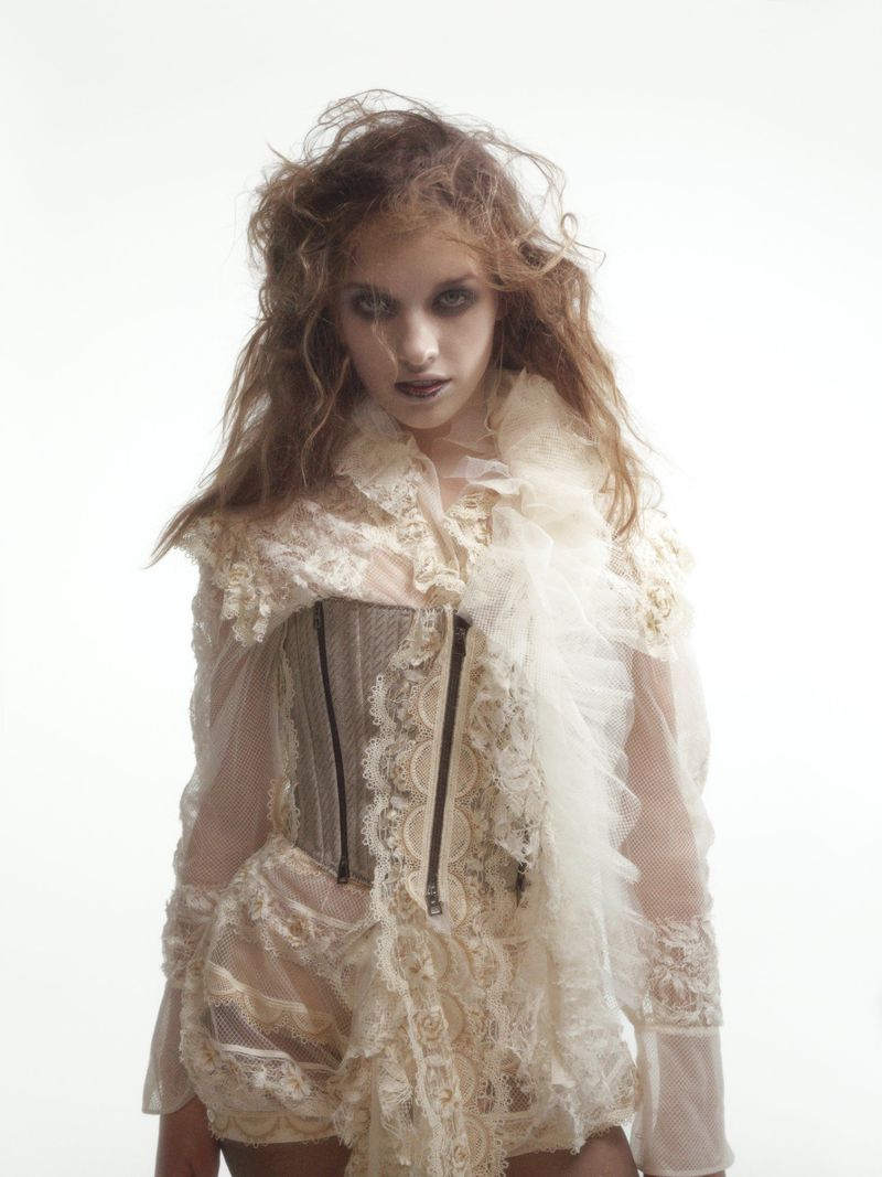 A young woman with dark eye makeup and ruffled white clothing stares into the camera against a bright white background.