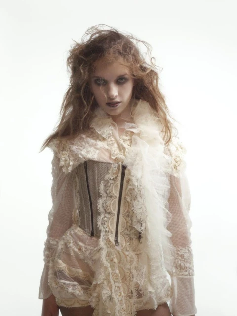A young woman with dark eye makeup and ruffled white clothing stares into the camera against a bright white background.