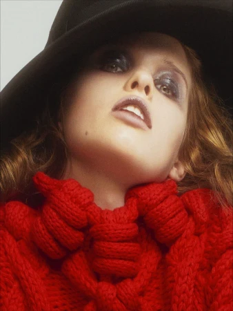 Ever Anderson wearing a red knit sweater and a wide black hat against a neutral backdrop in a portrait by Willy Vanderperre.