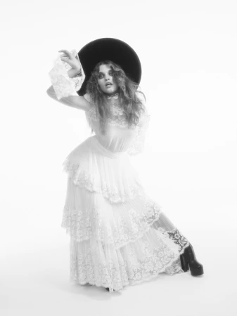 Ever Anderson in a tiered white lace dress and wide-brim black hat poses against a bright white seamless background.