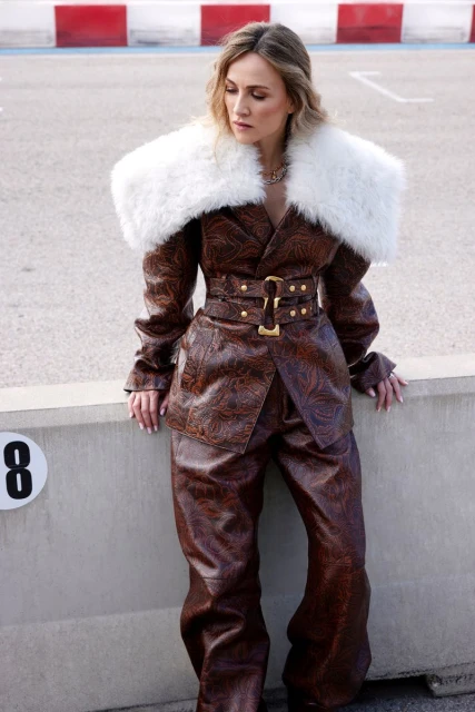 A woman poses outdoors on a racetrack wearing a brown leather suit with a white faux fur collar, exuding high fashion style.