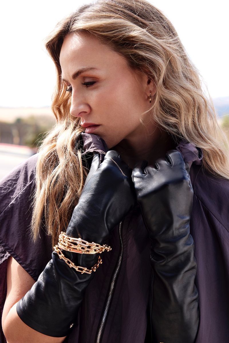 Woman in a fashionable oversized jacket, black leather gloves, and layered gold bracelets, posing outdoors in soft, natural light.