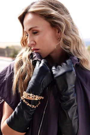 Woman in a fashionable oversized jacket, black leather gloves, and layered gold bracelets, posing outdoors in soft, natural light.