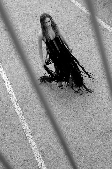 Black and white fashion shot of a woman in a fringe dress standing on asphalt, viewed from above, with metal bars in the foreground.