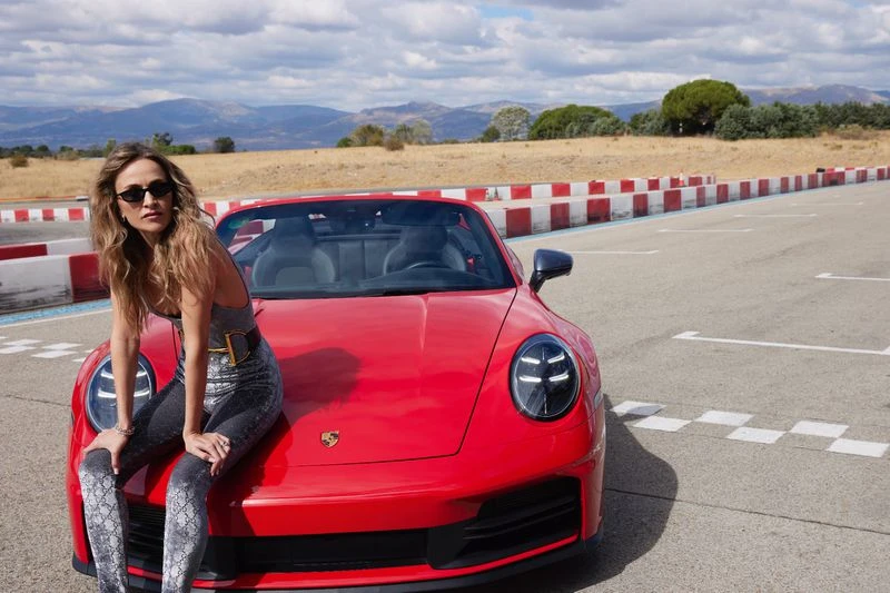 Woman poses on a red Porsche convertible on a racetrack with mountains in the background on a sunny day in a medium outdoor shot.