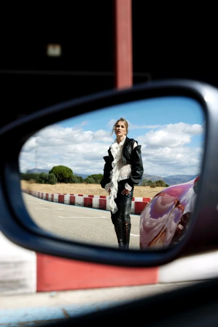 Reflection in a car mirror captures a woman in a black jacket and leather pants standing on a roadside with fields and sky.