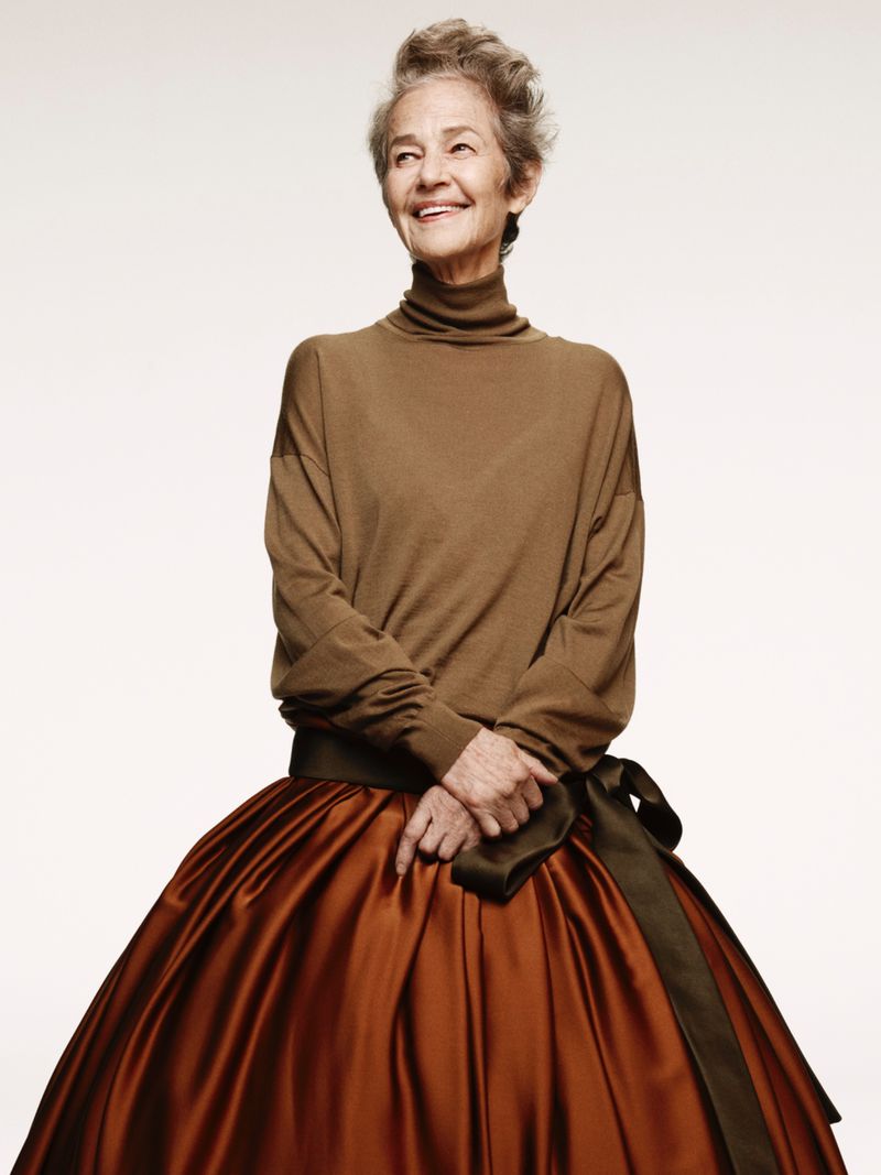 Elegant elderly woman smiles in a brown turtleneck and a large brown satin skirt with a bow against a clean, bright white background.