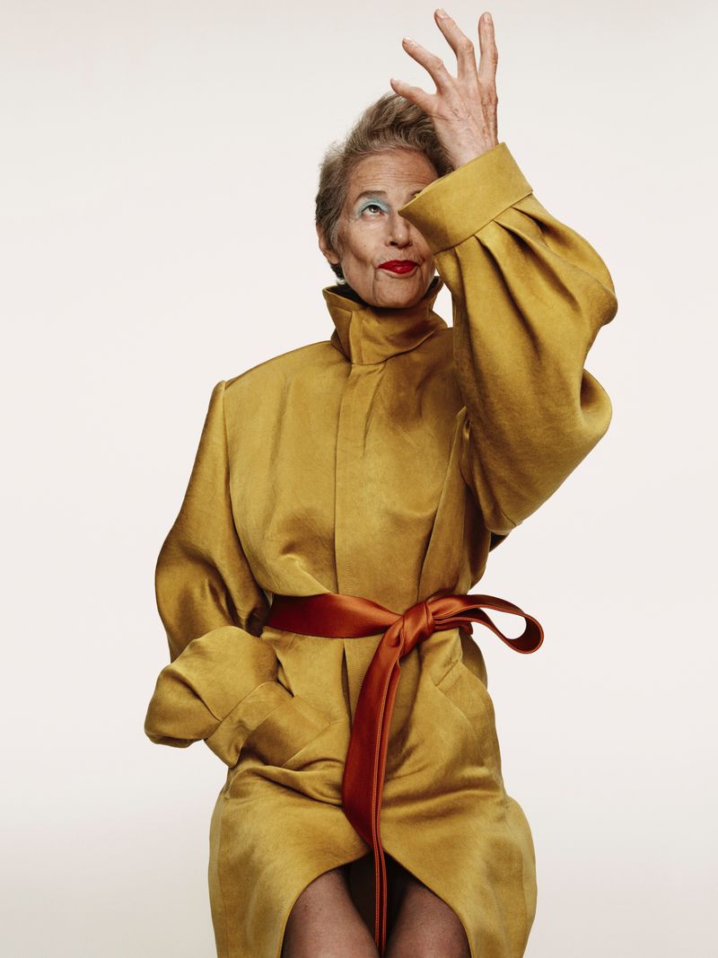 Elderly woman seated in a yellow coat with a red sash, hand raised near her face in a light studio setting for a fashion editorial.