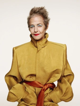 Elegant elderly woman in a golden coat with an orange sash poses in a studio setting with a neutral background.