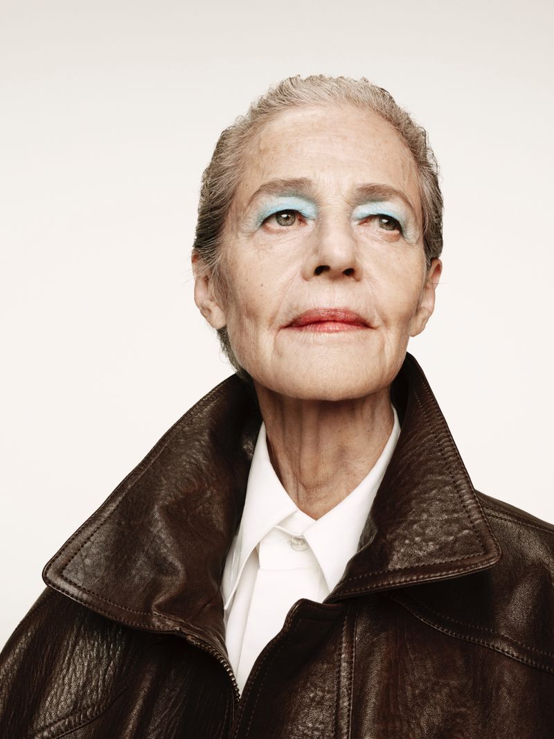 Close-up studio portrait of an elderly woman with blue eyeshadow wearing a brown leather jacket and a white collared shirt, looking up.
