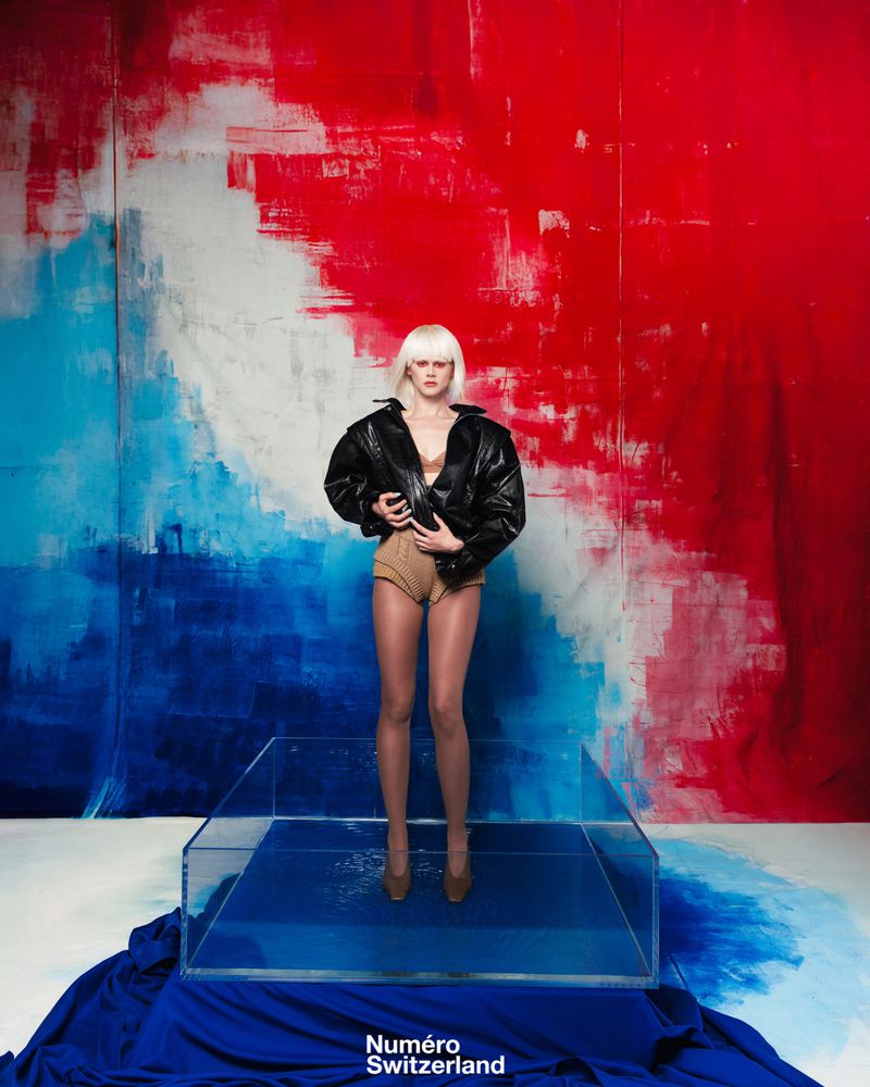 Fashion editorial shot of a woman with a blonde wig wearing a black jacket and high heels, standing in a blue water-filled glass box.