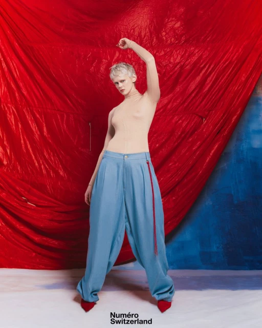 Fashion shot of a person with short white hair wearing blue pants and red high heels, striking a pose in front of a vibrant backdrop.