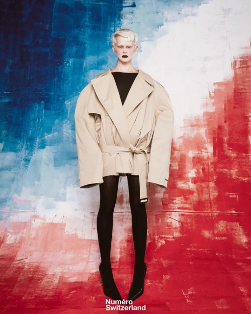Fashion shot of woman in oversized beige trench coat against a textured background painted with blue and red brush strokes.
