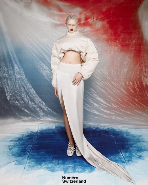Fashion editorial image of a young adult woman with platinum hair wearing a white outfit with sneakers in a studio environment.