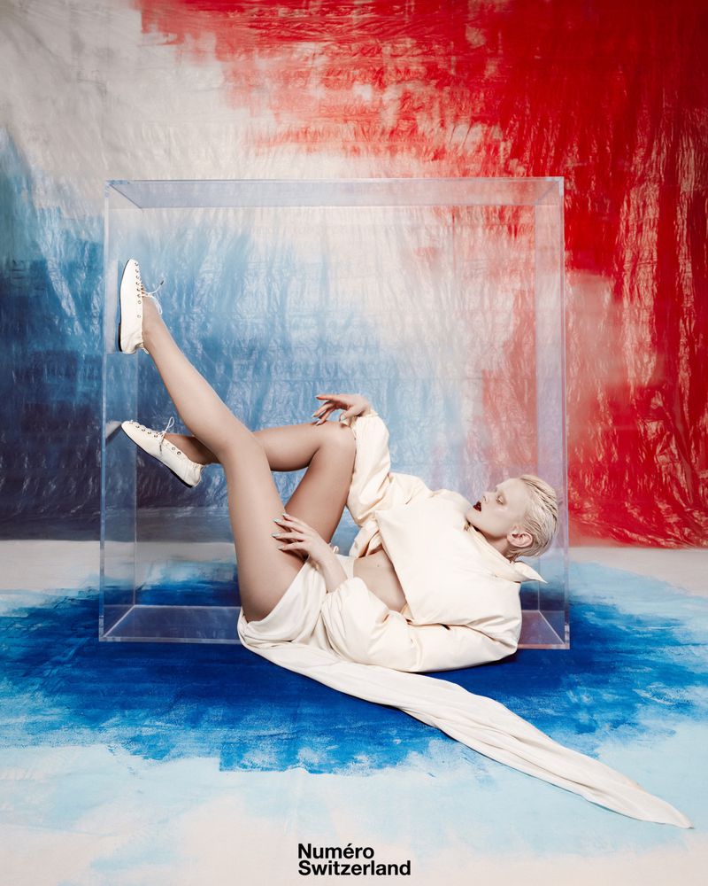Fashion model posing inside clear cube in front of red, white, and blue backdrop wearing a white jacket and sneakers in a studio.