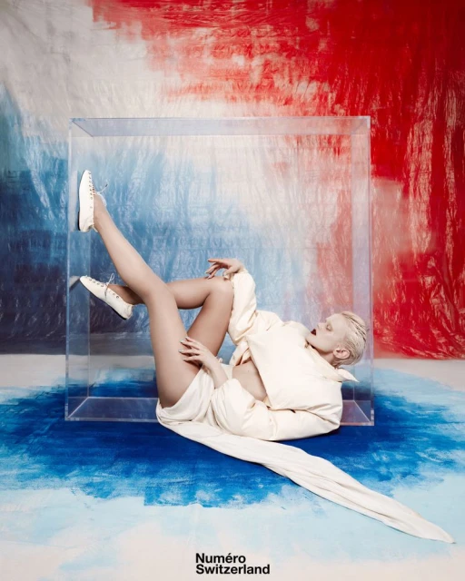 Fashion model posing inside clear cube in front of red, white, and blue backdrop wearing a white jacket and sneakers in a studio.