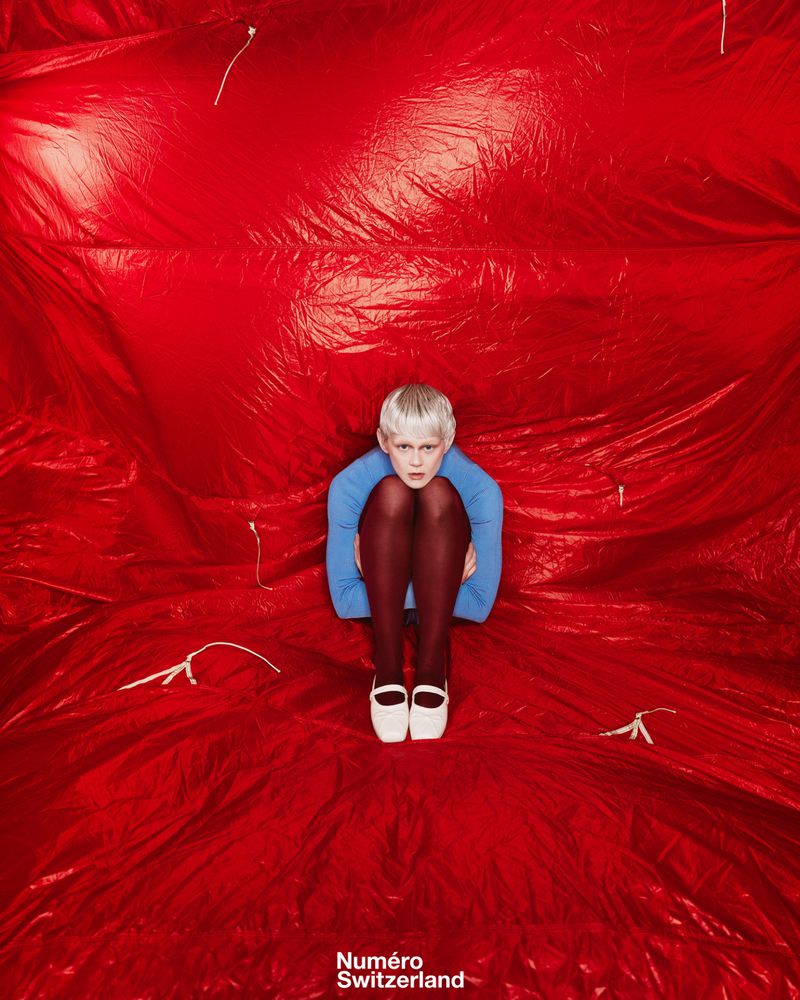 Child with short blond hair poses in blue top, brown tights and white shoes on a shiny red crumpled surface, looking at the camera.