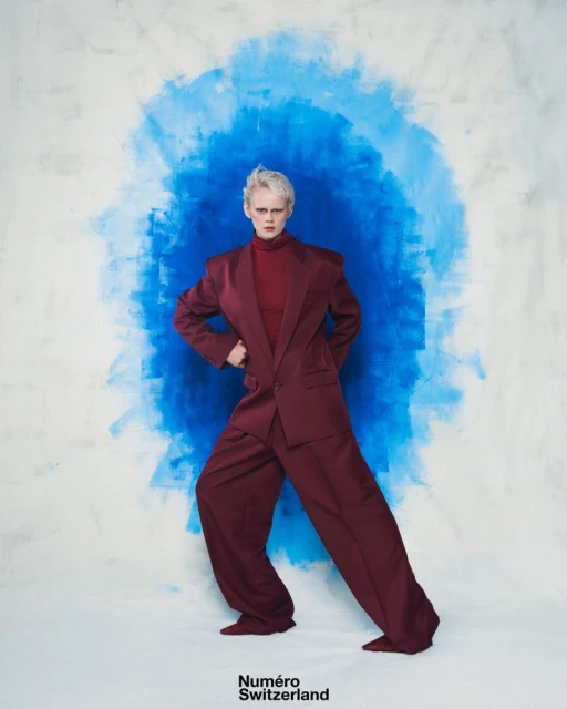 A female fashion model with short white hair poses in a maroon suit against a painterly blue and white backdrop in a studio shot.