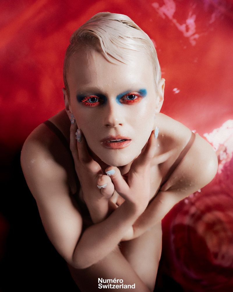 Close-up of fair-skinned person with artistic makeup, posing with hands near face against red backdrop and wearing a minimal shoulder strap.