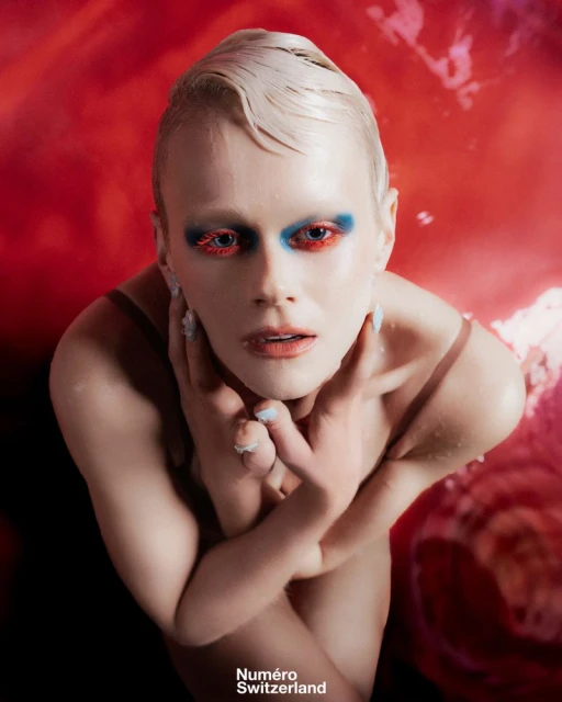 Close-up of fair-skinned person with artistic makeup, posing with hands near face against red backdrop and wearing a minimal shoulder strap.