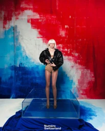 Model with platinum blonde hair wearing a black jacket and nude briefs standing inside a clear acrylic box against a red and blue painted backdrop.