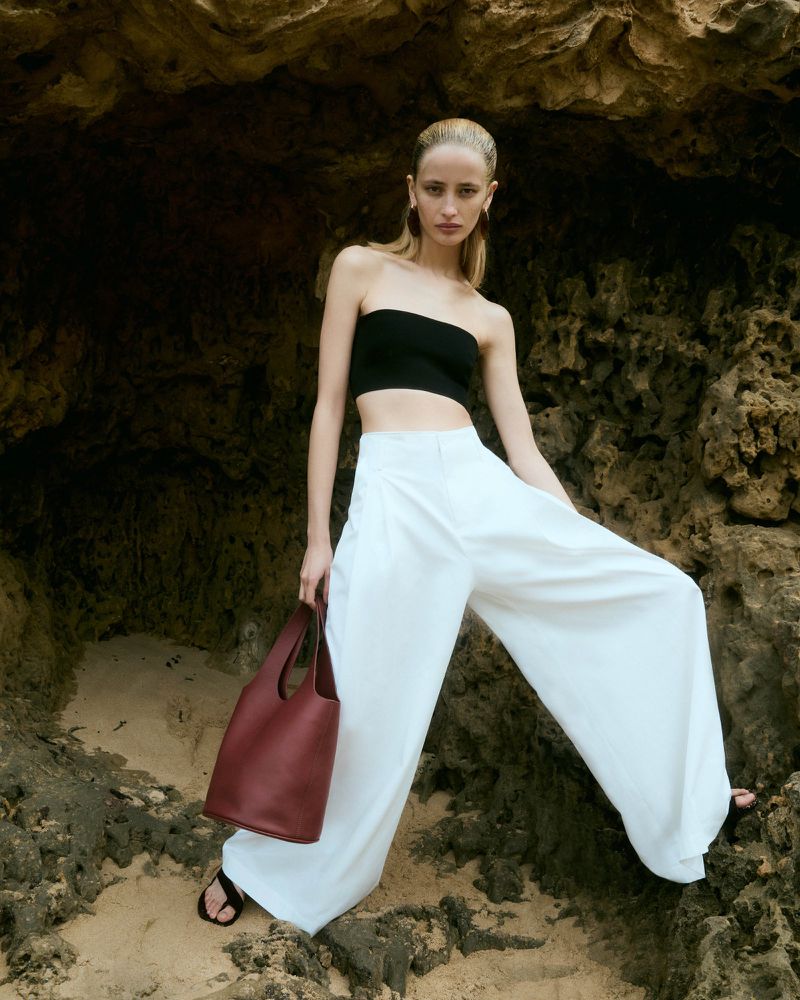 Young woman wearing a black tube top and white wide-leg pants, posing with a red leather tote bag in a beachy rocky environment.