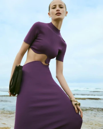 Model Nabila Leunig in a purple short-sleeved cutout dress holding a black clutch bag by the ocean shore.