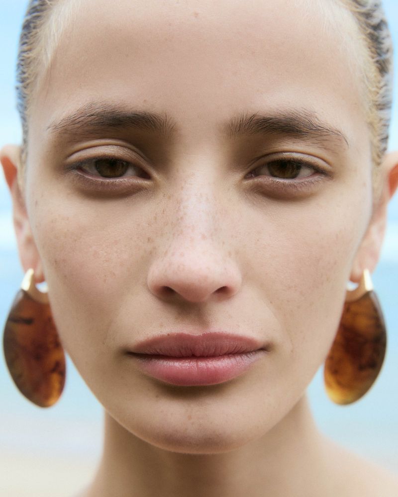 Close-up portrait of a young adult woman with brown hair, fair skin, and freckles, wearing brown earrings, with an outdoor background.