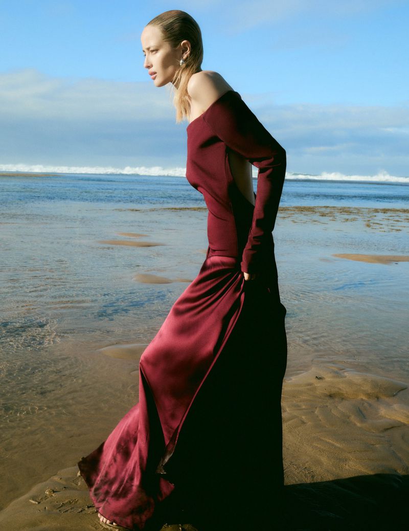 A woman in a long, dark red dress stands on a sandy beach next to the ocean. The sky is blue with some clouds. Fashion photography.
