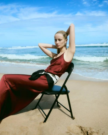 Model Nabila Leunig wearing a long burgundy satin dress while seated on a folding chair on a sandy beach by the ocean.
