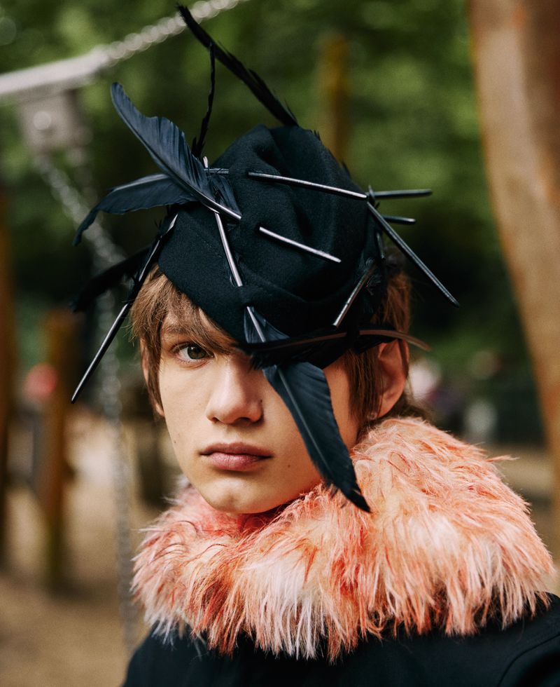 Portrait of an adult male with a black hat decorated with feathers and sticks, wearing a fuzzy peach-colored collar and a black jacket.