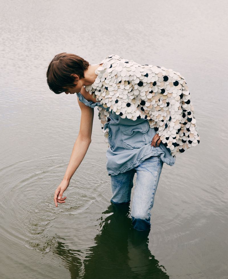 Woman in a flowing, circular-patterned top and jeans stands in shallow water, gently touching the surface with her hand, creating ripples.