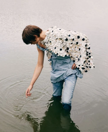 Woman in a flowing, circular-patterned top and jeans stands in shallow water, gently touching the surface with her hand, creating ripples.