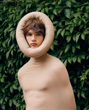 A person with fair skin and light brown hair is wrapped in nude fabric against green foliage in a portrait-style, eye-level shot.