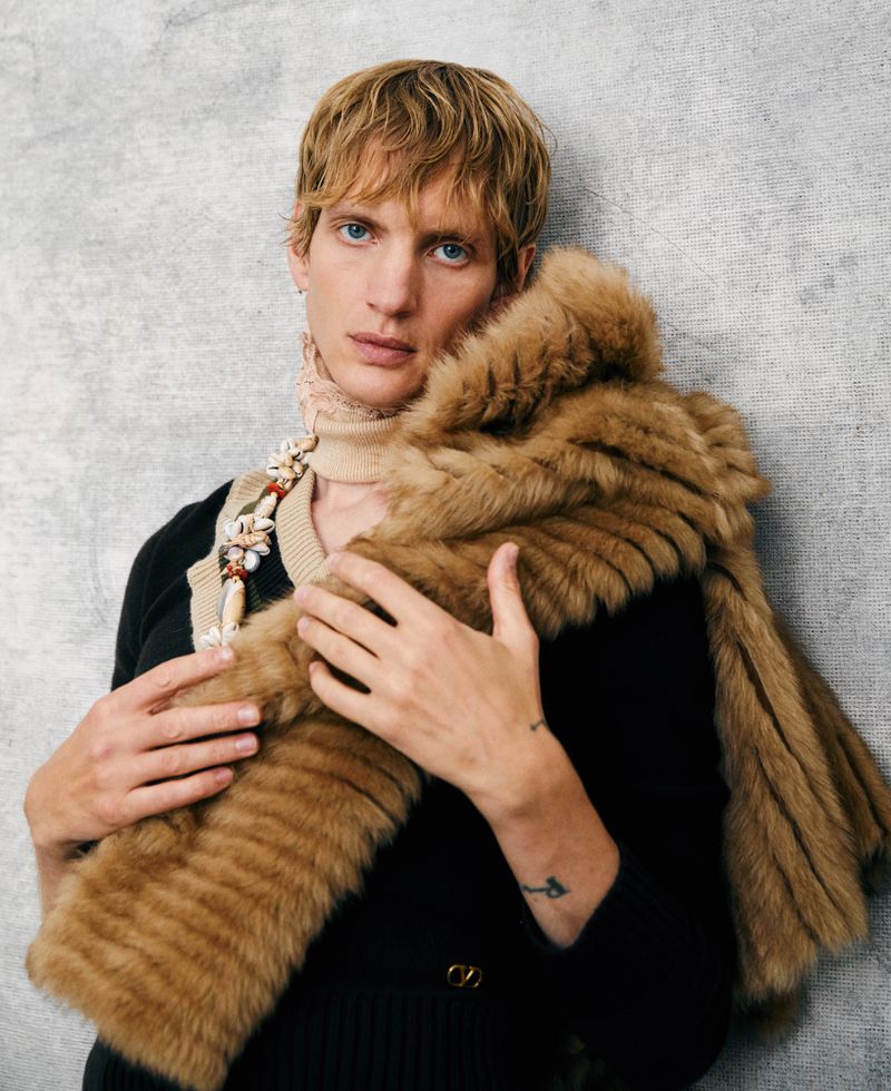 A fair-skinned young adult male with blond hair and blue eyes is styled with a brown fur stole indoors against a grey textured wall.