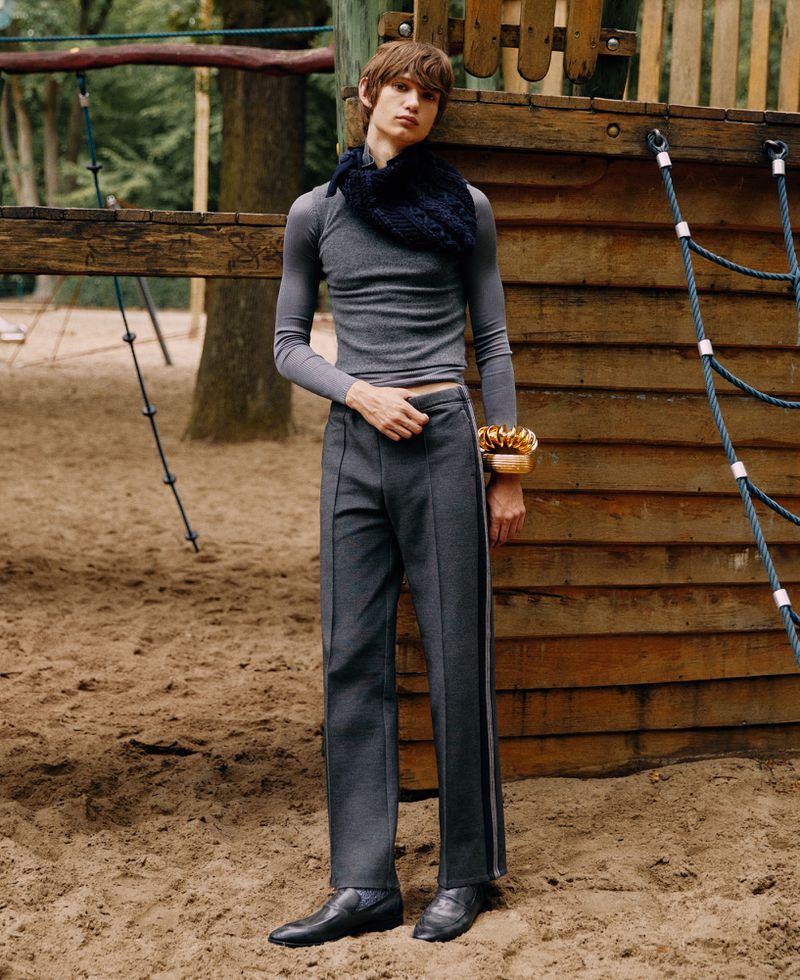 A slim adult male model in gray top and pants stands in outdoor fashion shot with playground, trees, and sandy ground.