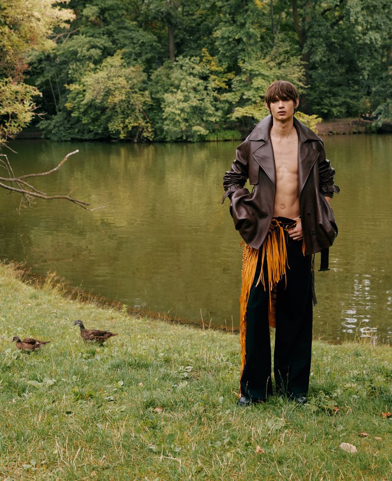 Adult male posing in leather jacket and fringed pants near lake, with ducks on grassy bank and forested background.