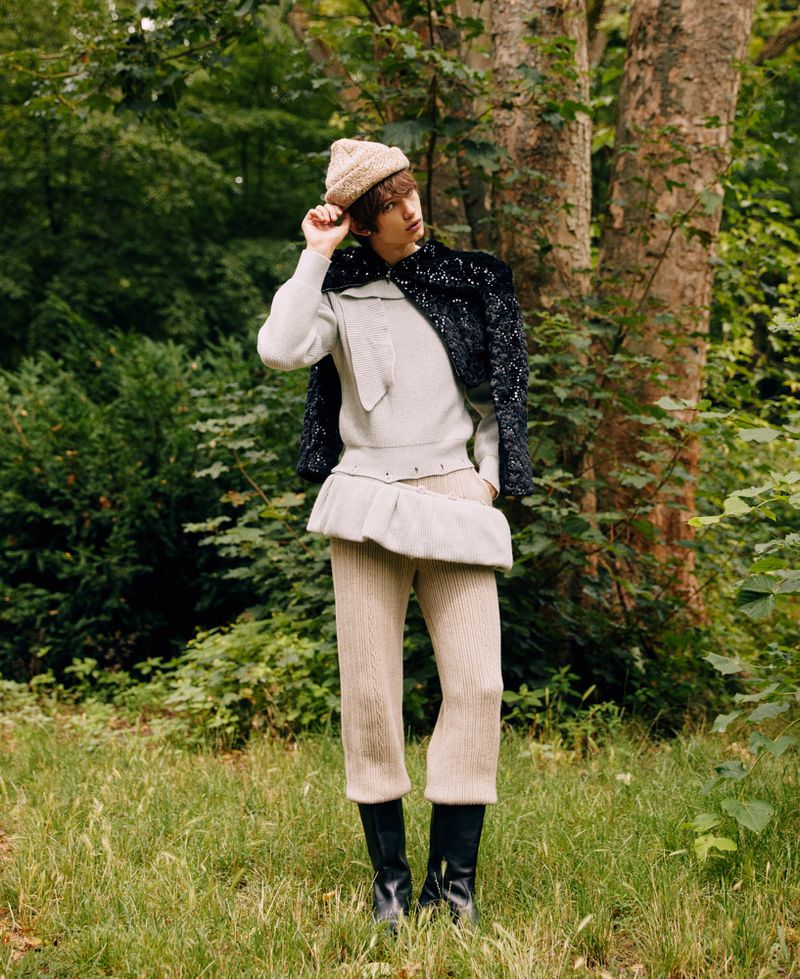 A person in a beige knit outfit and black boots poses in a forest, wearing a knit hat and a black textured jacket.