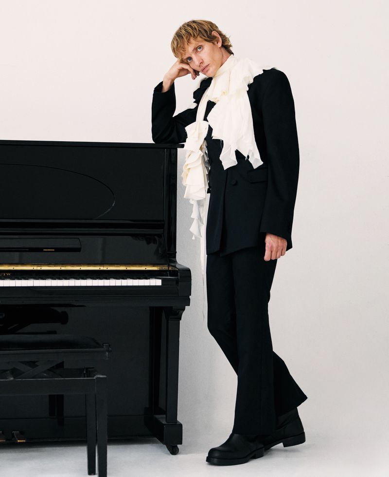 Young adult poses next to a black upright piano in a photo studio, wearing a dark suit with a ruffled white scarf.
