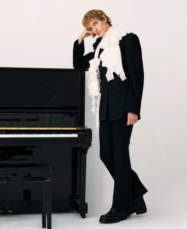 Young adult poses next to a black upright piano in a photo studio, wearing a dark suit with a ruffled white scarf.