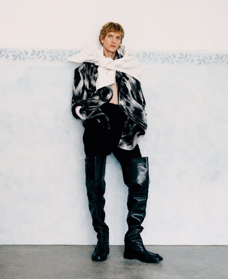 Model wearing a black and white patterned jacket with thigh-high black boots, leaning against a pale blue wall.