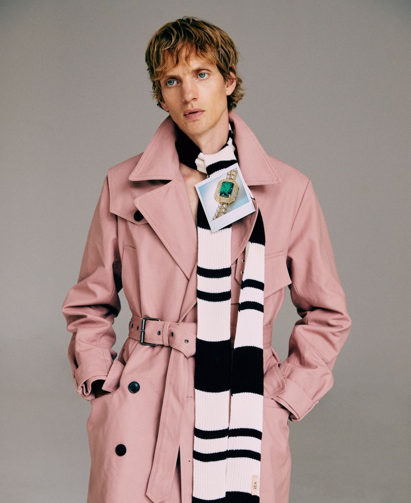 Young adult male with fair skin and blond hair posing indoors in a pink trench coat with a striped scarf in a fashion setting.