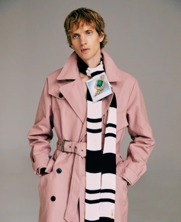 Young adult male with fair skin and blond hair posing indoors in a pink trench coat with a striped scarf in a fashion setting.